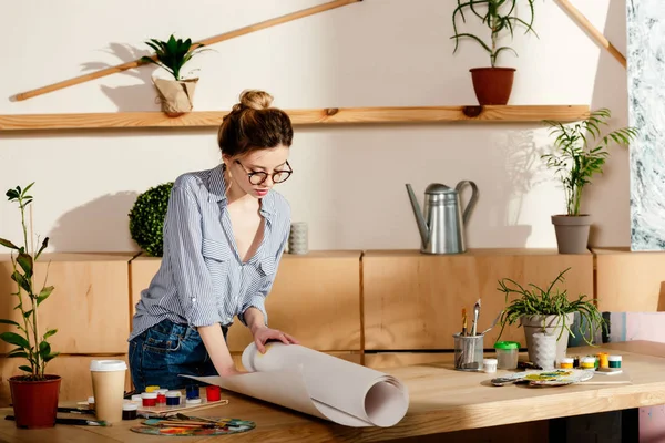 Attraente donna elegante in occhiali rotolamento tela sul tavolo con forniture pittura — Foto stock