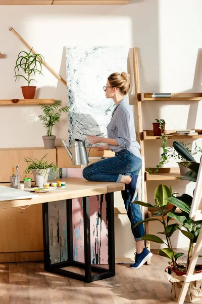 Side view of stylish female artist in eyeglasses watering potted plan in studio — Stock Photo