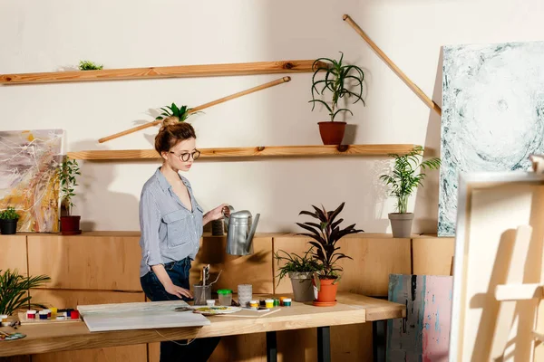 Young stylish female artist in eyeglasses watering potted plan in studio — Stock Photo