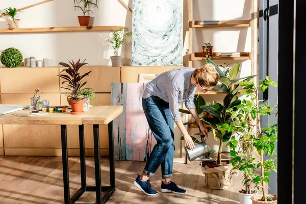 Stylish female artist in eyeglasses watering potted plan in studio — Stock Photo