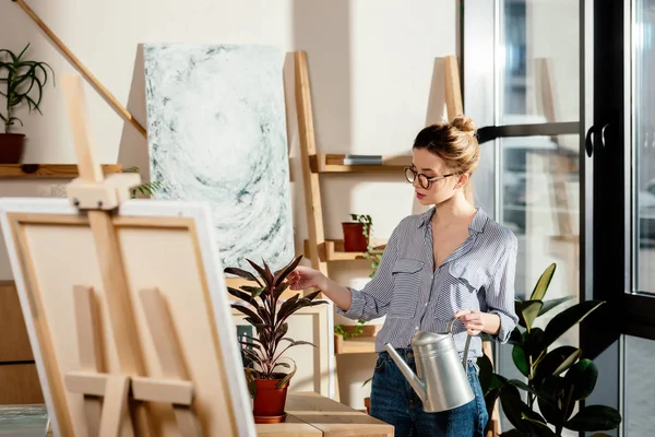 Attractive stylish female artist in eyeglasses holding watering can and touching plant — Stock Photo