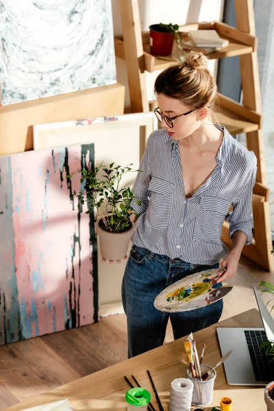 Élégant artiste féminine dans les lunettes tenant plante et palette en studio — Photo de stock