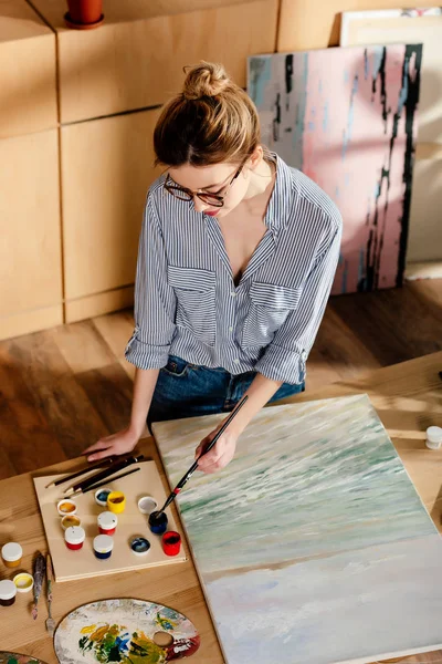 Vista de ángulo alto de la artista femenina con estilo en gafas poner pincel en la pintura - foto de stock