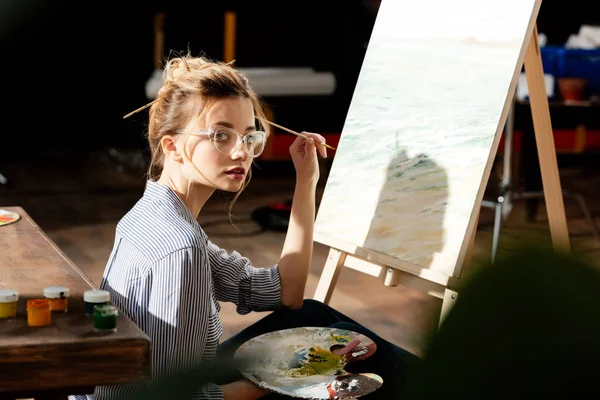 Молодая стильная художница в очках держит палитру и кисть — Stock Photo