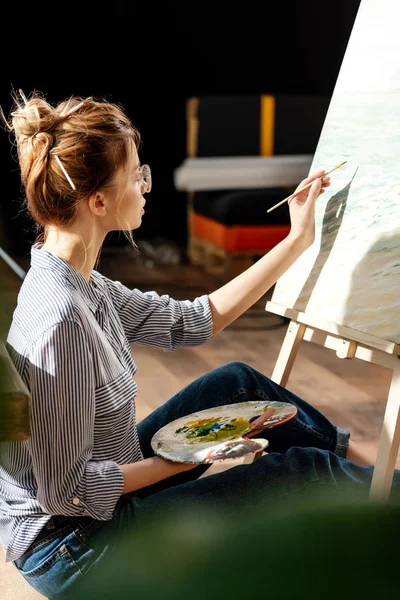 Side view of stylish female artist in eyeglasses  painting on easel — Stock Photo