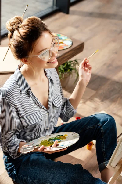 Smiling stylish female artist in eyeglasses holding palette and paintbrush — Stock Photo