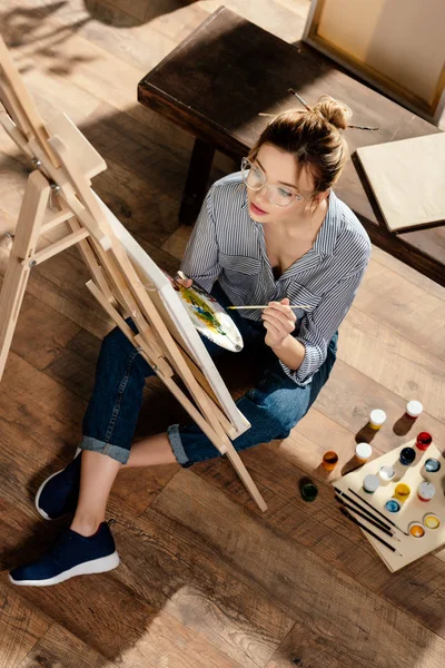 Vista de alto ângulo de artista feminina elegante na pintura de óculos no cavalete — Fotografia de Stock