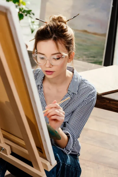 Young stylish female artist in eyeglasses painting on easel — Stock Photo