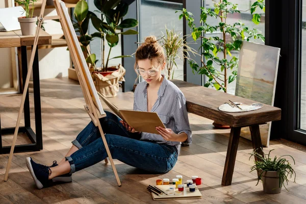 Elegante joven artista femenina en gafas de lectura libro y sentado en el suelo con materiales de pintura - foto de stock