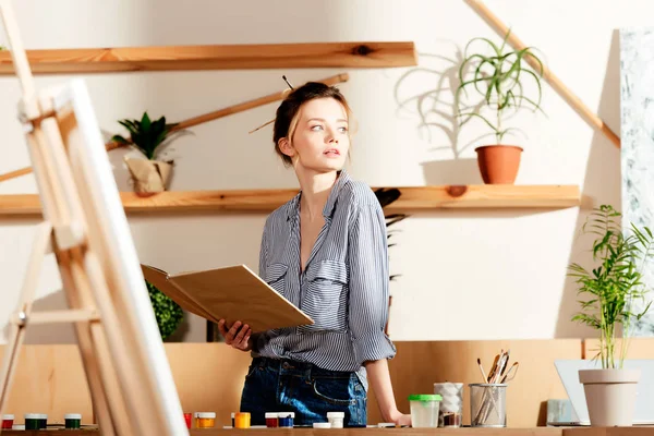 Jovem artista feminina atraente segurando livro e olhando para longe — Fotografia de Stock