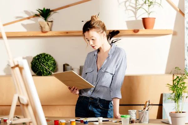 Young female artist reading book and standing at table with painting supplies — Stock Photo