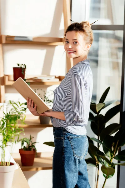 Sourire jeune femme lecture livre près de plantes en pot — Stock Photo