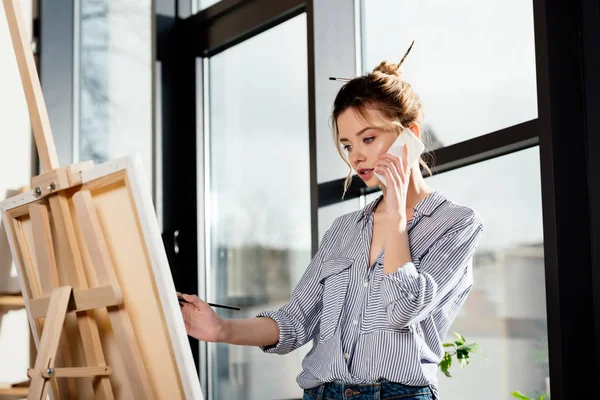 Young female artist talking on smartphone and painting on easel — Stock Photo