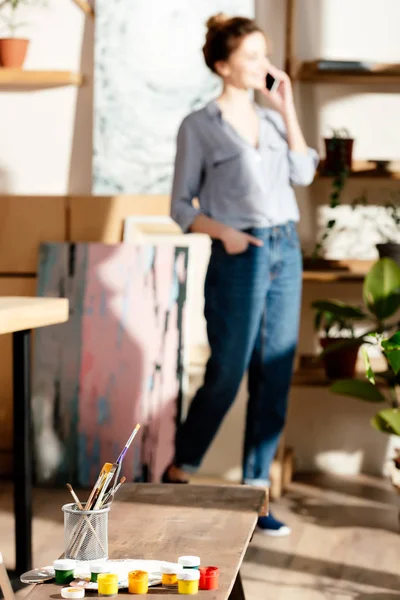 Materiales de pintura en el banco y artista femenina hablando en el teléfono inteligente detrás - foto de stock