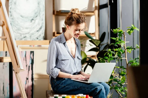Усміхнена молода жінка друкує на ноутбуці і сидить на лавці з фарбами — стокове фото