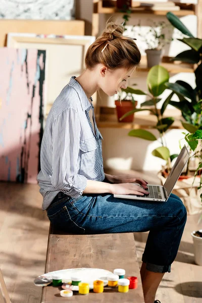 Vue latérale de la jeune artiste féminine assise sur un banc avec des peintures et utilisant un ordinateur portable — Photo de stock