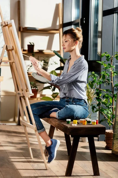 Side view of female artist sitting on bench with paints and drawing picture on easel — Stock Photo