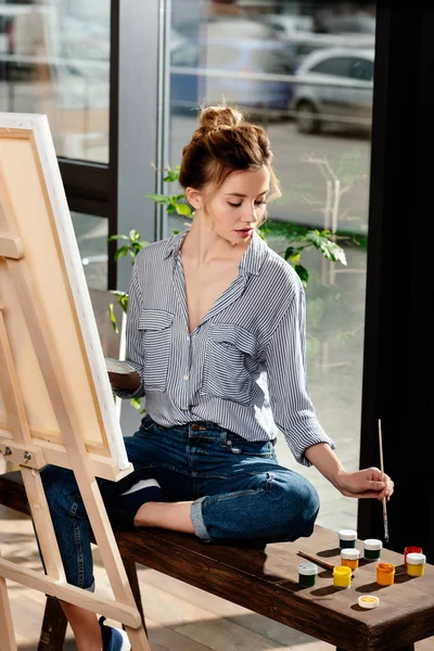 Female artist sitting on bench with paints and drawing picture on easel — Stock Photo