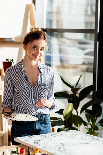 Portrait of young female artist with palette and paintbrush in art studio — Stock Photo