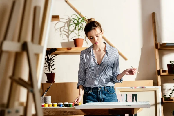 Young female artist standing with paintbrush and looking at painting — Stock Photo