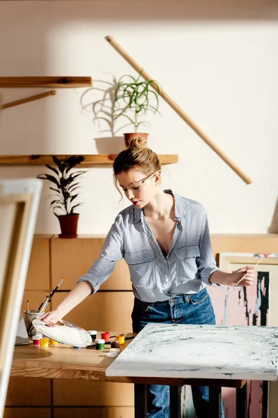 Artista femenina con pincel detrás de la oreja tomando paleta en la mesa con pintura y pinturas - foto de stock