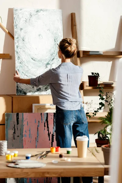 Rear view of female artist with paintbrush behind ear taking off canvas in studio — Stock Photo