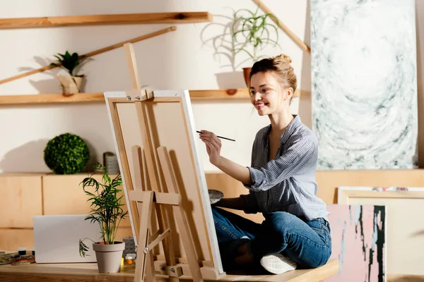 Sonriente artista sentada en la mesa y dibujando sobre lienzo en el estudio - foto de stock