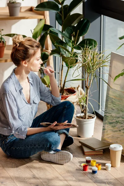 Side view of young female with paintbrush and palette sitting at floor in studio — Stock Photo