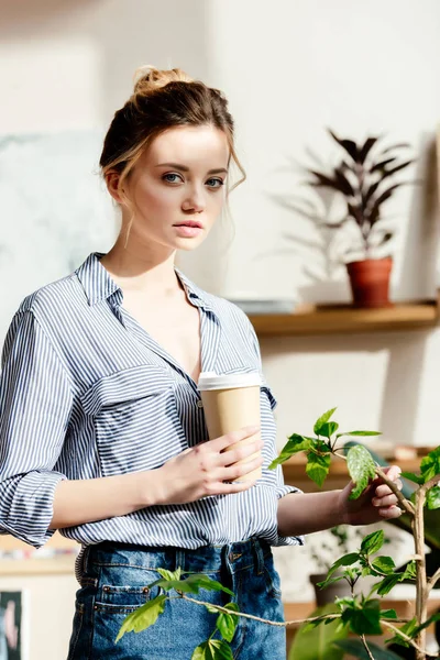 Ritratto di giovane donna con tazza di carta di caffè toccante pianta in vaso — Foto stock