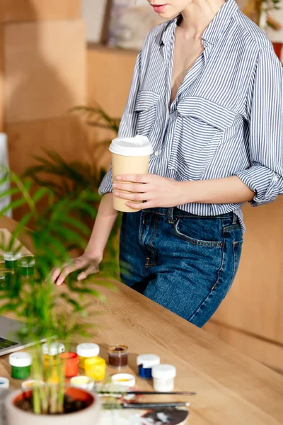 Colpo ritagliato di giovane donna in possesso di tazza di carta di caffè a tavola con computer portatile e vernici — Foto stock