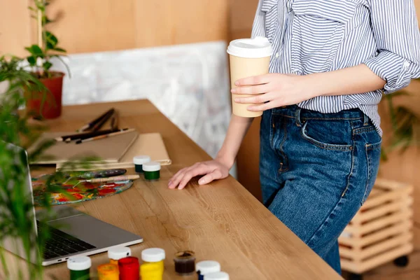 Cropped image of female artist with coffee cup standing at table with laptop and painting supplies — Stock Photo