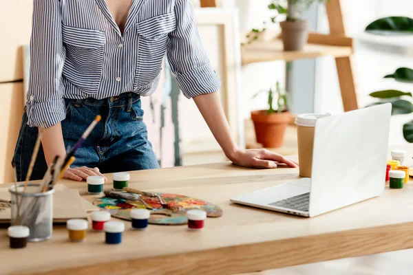 Tiro recortado de artista feminina em pé à mesa com laptop e materiais de pintura — Fotografia de Stock