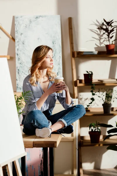 Carta vuota su cavalletto e artista donna seduta sul tavolo con tazza di caffè usa e getta — Foto stock