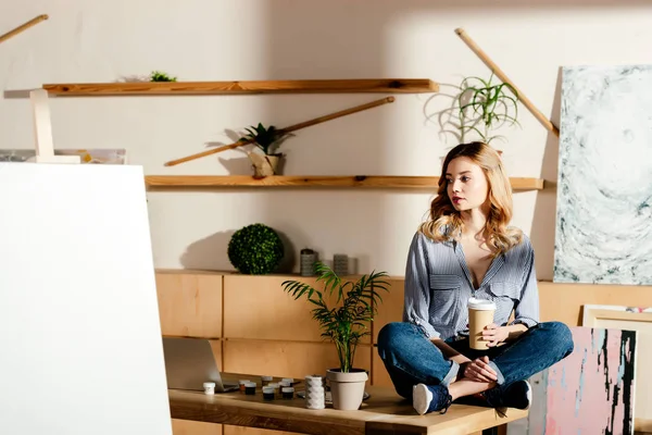 Jeune artiste féminine avec tasse de café en papier assis sur la table en studio — Photo de stock