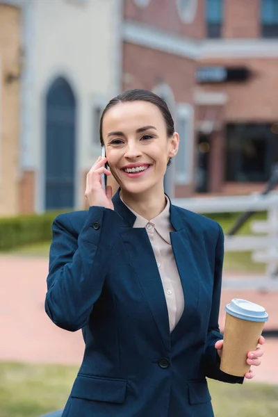 Sonriente hermosa mujer de negocios hablando por teléfono inteligente y la celebración de café en taza de papel - foto de stock