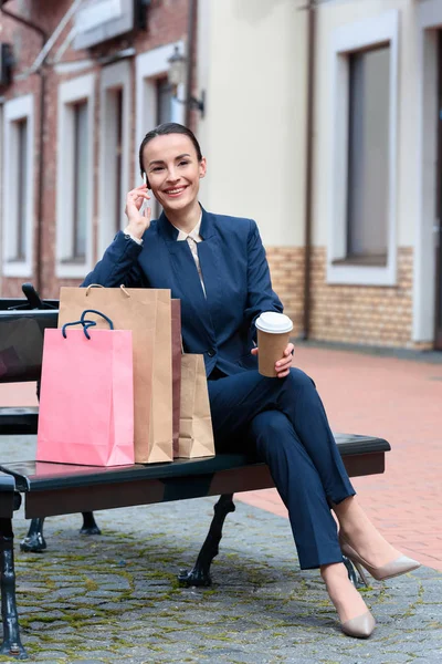 Attraktive Geschäftsfrau sitzt mit Einkaufstaschen und Kaffee auf Bank und unterhält sich per Smartphone — Stockfoto