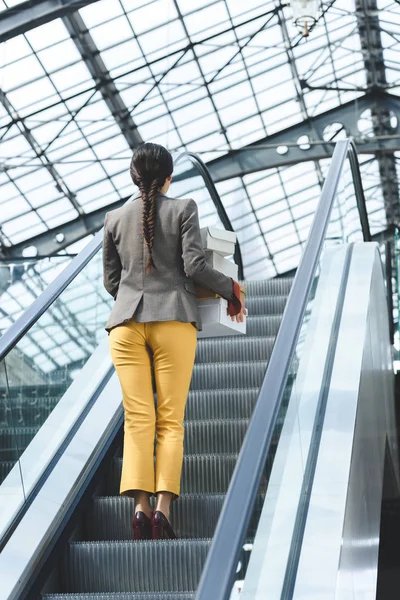 Vista trasera de la mujer de pie con cajas de compras en la escalera mecánica en el centro comercial - foto de stock
