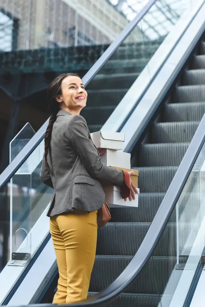 Happy attractive woman standing on escalator in mall with shopping boxes — Stock Photo