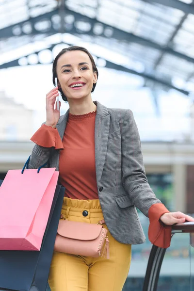 Attraente donna parlando da smartphone e tenendo borse della spesa nel centro commerciale — Foto stock