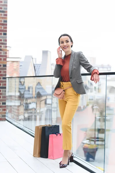 Donna in piedi sul balcone del centro commerciale con borse della spesa vicino alle gambe e parlando con lo smartphone — Foto stock