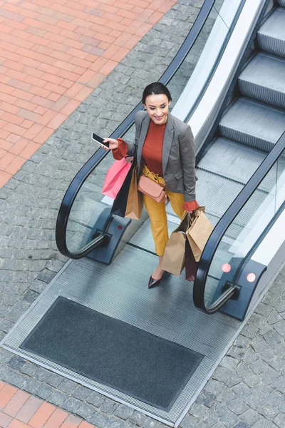 Vue grand angle de la femme attrayante marchant avec des sacs à provisions de l'escalator dans le centre commercial — Photo de stock