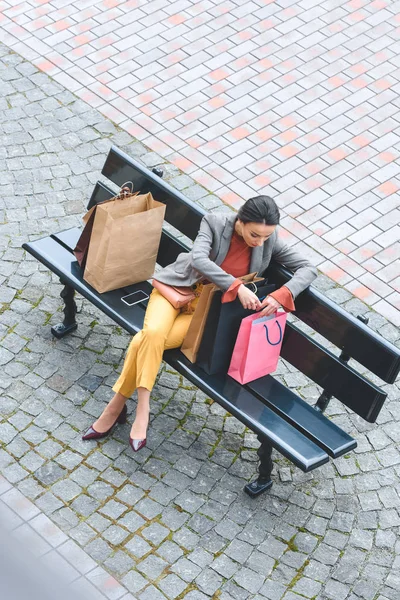 Vue grand angle de la femme attrayante assise sur le banc et regardant dans le sac à provisions — Photo de stock