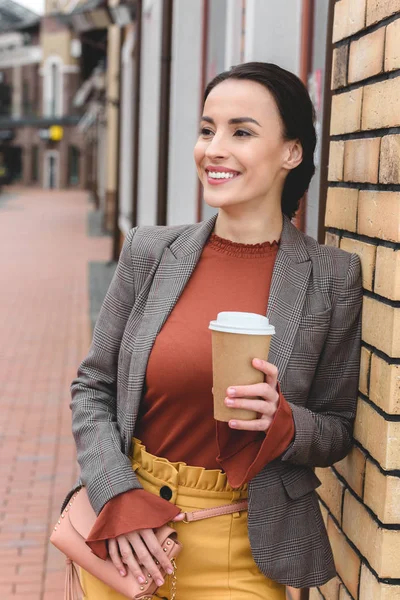 Feliz bela mulher elegante segurando café no copo de papel e inclinando-se na parede — Fotografia de Stock