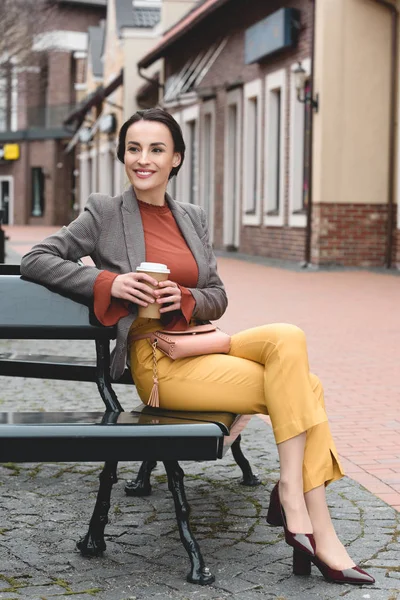 Sorridente bella donna elegante seduta su panchina con caffè in tazza di carta — Foto stock