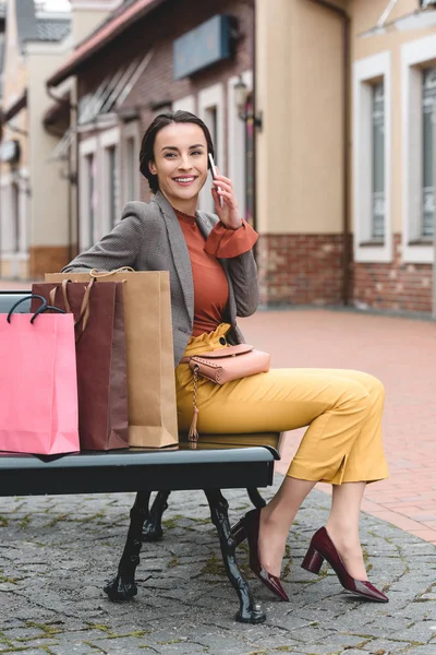 Schöne Frau sitzt mit Einkaufstüten auf Bank und unterhält sich per Smartphone — Stockfoto