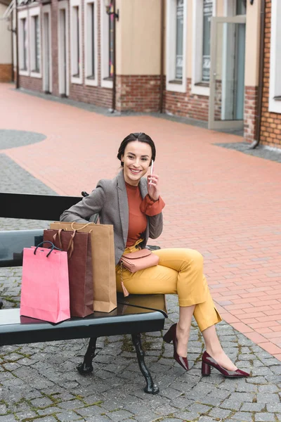 Attraktive Frau sitzt mit Einkaufstüten auf Bank und unterhält sich per Smartphone — Stockfoto