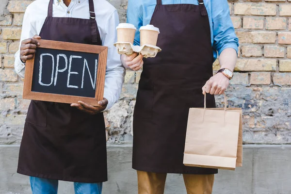 Tiro recortado de dos propietarios multiétnicos de la cafetería sosteniendo cartel abierto, bolsas de papel y tazas de café desechables mientras está de pie cerca de la pared de ladrillo - foto de stock