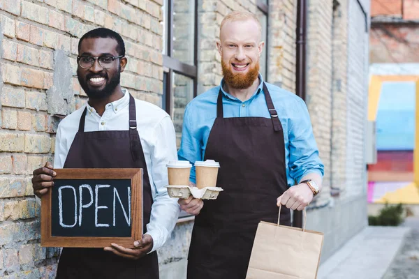 Young handsome multiethnic owners of coffee shop in aprons with sign open, paper bags and disposable coffee cups smiling at camera — Stock Photo