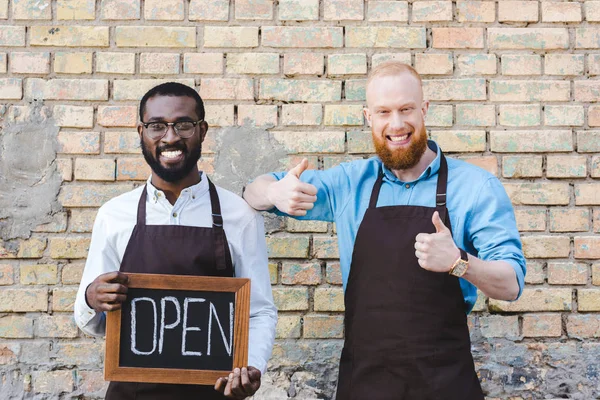 Gutaussehende multiethnische Coffeeshop-Besitzer in Schürzen halten Schild offen und zeigen Daumen hoch, lächeln in die Kamera — Stockfoto