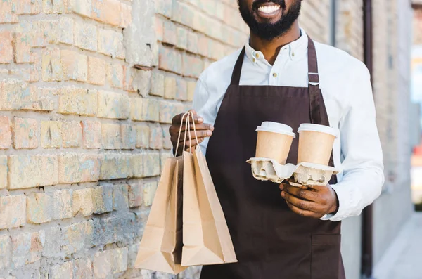 Recortado disparo de sonriente joven africano americano barista sosteniendo bolsas de papel y tazas de café desechables - foto de stock